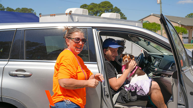A volunteer stands next to a pet owner holding a small dog in his car, both are smiling