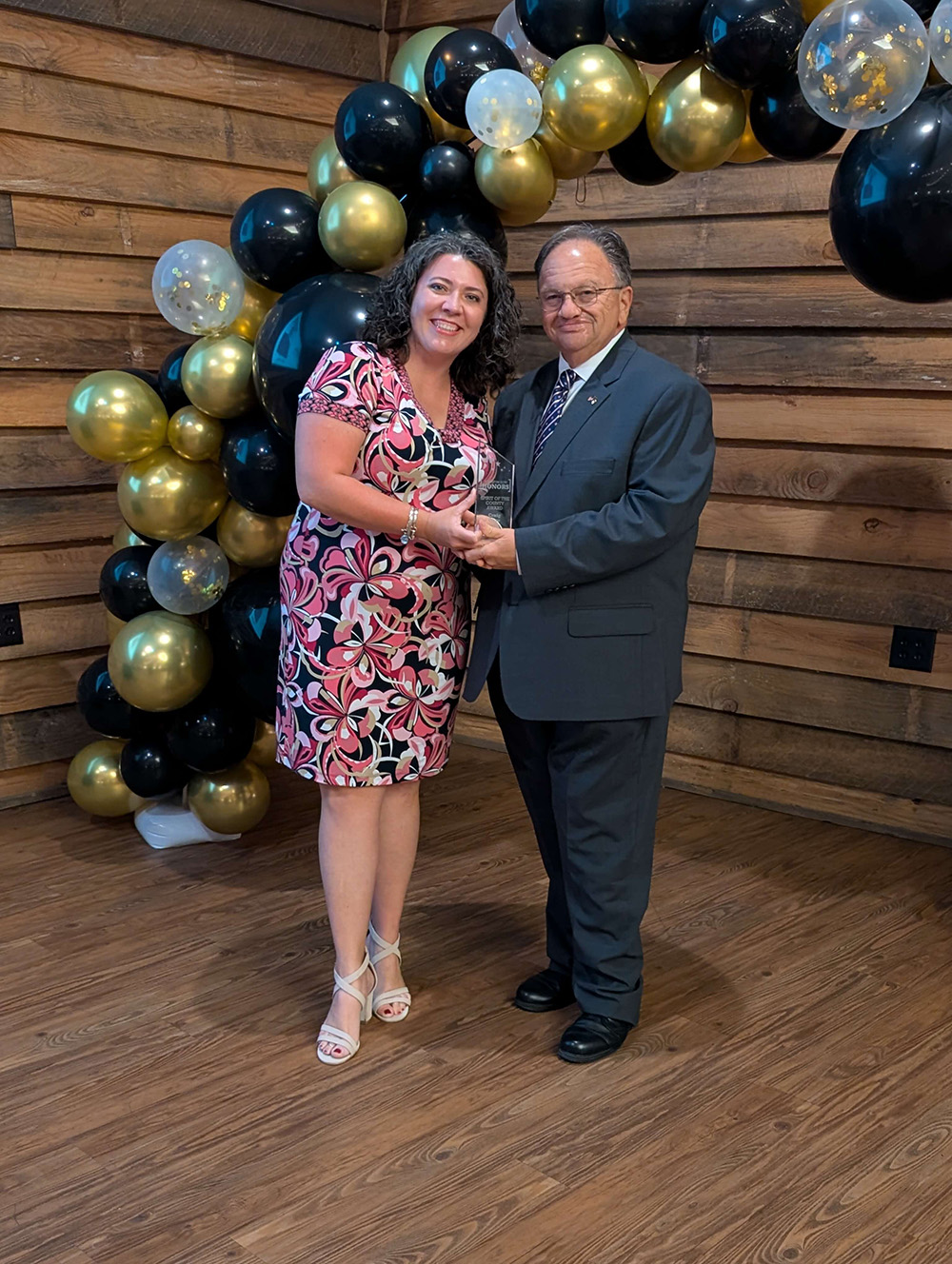 Photo of Craig Olive and a woman both holding a glass award with balloon arch in background